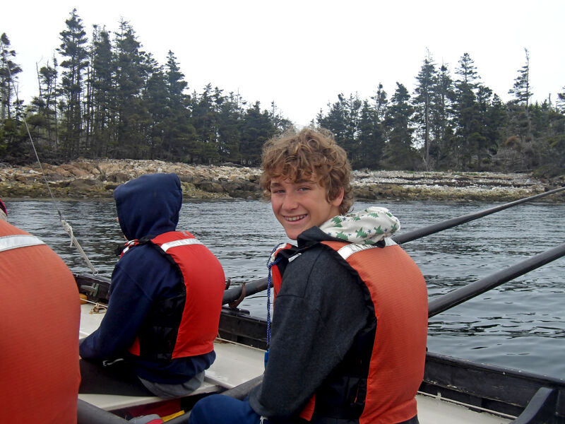 The image shows a group of people in a boat, likely rowing. They are wearing life vests. One person is looking at the camera and smiling. The background includes trees and water, suggesting they are on a lake or the ocean. The weather appears overcast.
